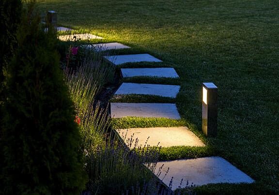 marble path of square tiles illuminated by a lantern glowing with a warm light in a backyard garden with a flower bed and a lawn copy space.